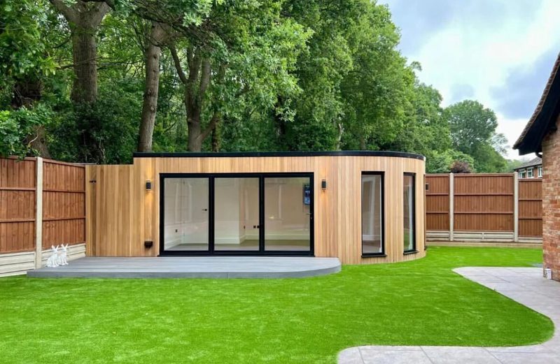 Curved wooden garden room with black-framed glass doors on a curved patio, fenced yard, green lawn and tall trees behind.