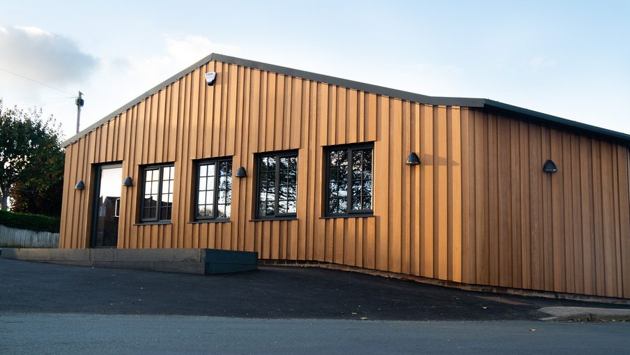 Single-story building with vertical wooden siding in warm brown, black-framed windows, exterior lights, a small ramp, and a paved parking area in front under a gabled roof.