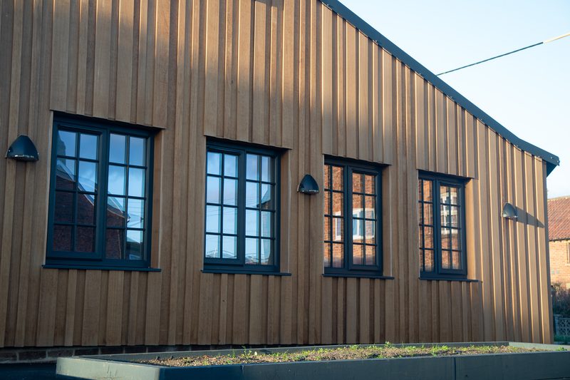 Brown vertical wooden-clad building with four dark-framed windows, wall-mounted lights, and a raised planter bed in front under a clear blue sky.