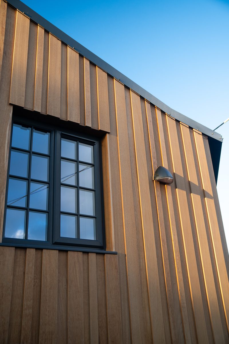 Modern wooden exterior with vertical brown panels, a black-framed grid window, and a wall light against a clear blue sky.