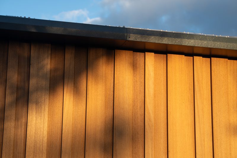 Sunlit vertical wooden wall panels in warm amber tones, with a dark metal roof edge at the top; shadows on the planks and a blue sky with clouds.
