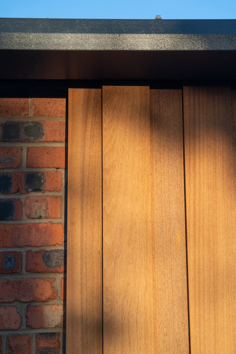 Close-up of a red brick wall beside vertical wooden panels under a dark overhang, with blue sky above and sunlight casting shadows on the wood.