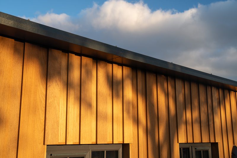Sunlit wooden wall with vertical planks, warm glow and branch shadows, a dark metal roof edge, and a small window at the bottom against a blue sky.