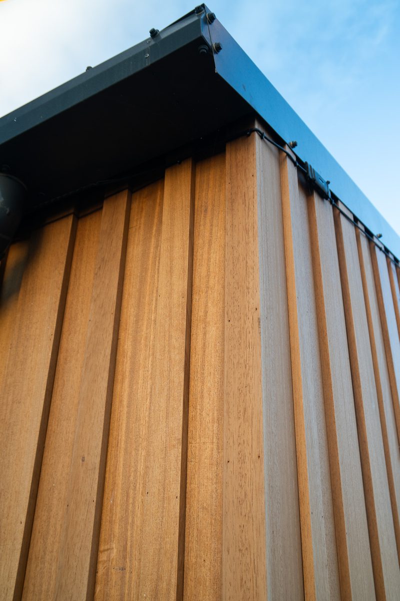 Low-angle view of a wooden exterior wall with vertical brown planks, a dark metal roof edge with bolts, and a blue sky.