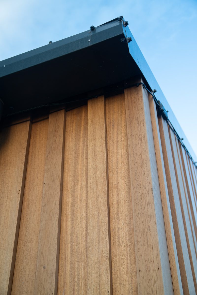 Close-up of vertical wooden siding under a dark metal roof edge, with visible screws, against a clear blue sky.