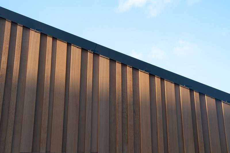 Diagonal view of a brown wooden slat wall with vertical panels and a dark top trim, against a clear blue sky.