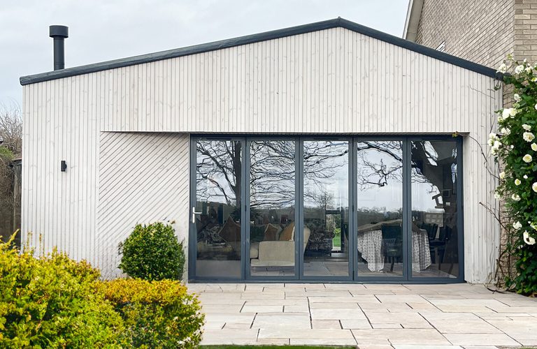 Modern single-story extension with large glass doors, light wood cladding, and a patio; green lawn, shrubs, and climbing white flowers on the right.