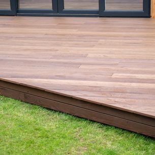 Brown wooden deck with evenly spaced planks, raised above green grass; dark-framed glass doors are visible at the rear.