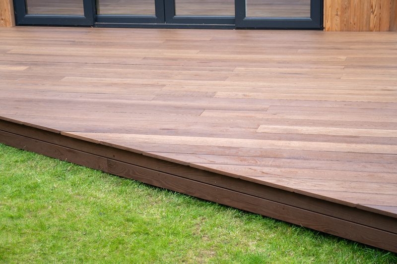 Brown wooden deck with evenly spaced planks, raised above green grass; dark-framed glass doors are visible at the rear.