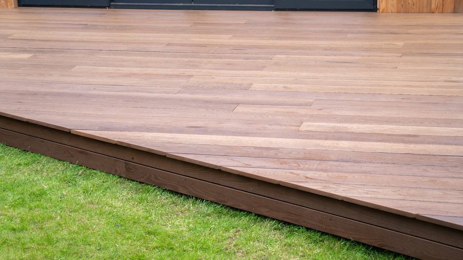 Brown wooden deck with evenly spaced planks, raised above green grass; dark-framed glass doors are visible at the rear.