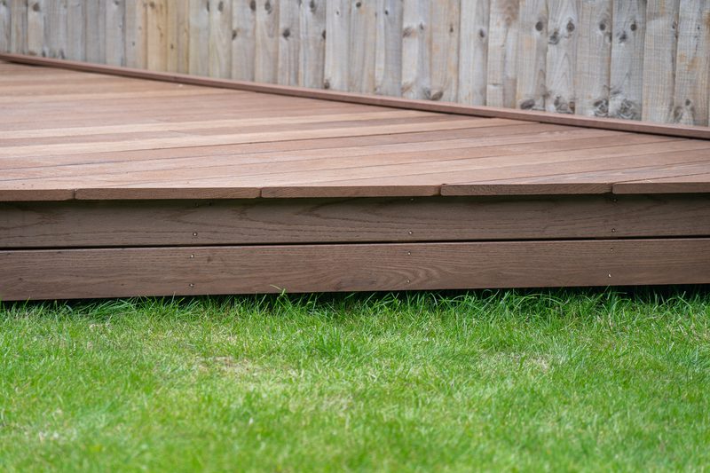 Elevated brown wooden deck with visible plank seams over a green lawn, backed by a wooden fence.