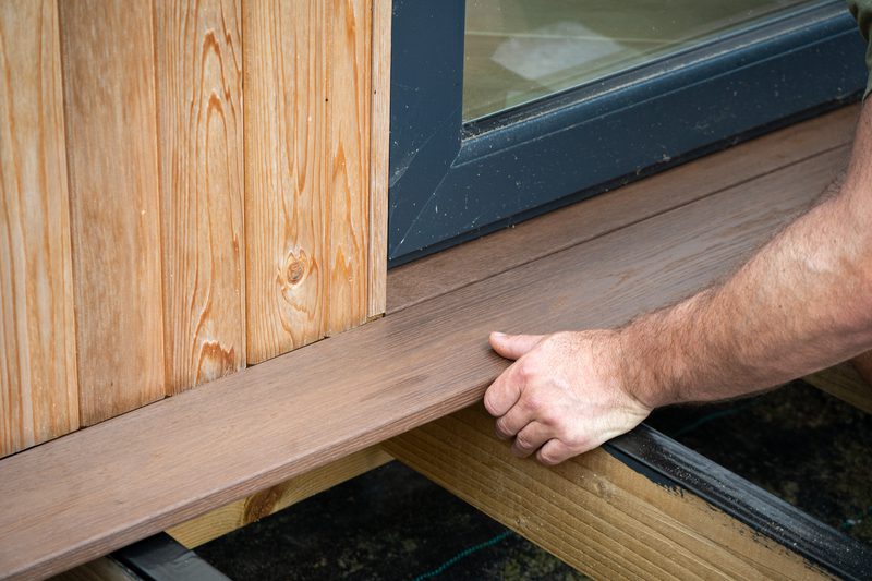 Close-up of a hand resting on a wooden deck plank beside a glass door, with vertical wood siding on the left and a blue-framed window.