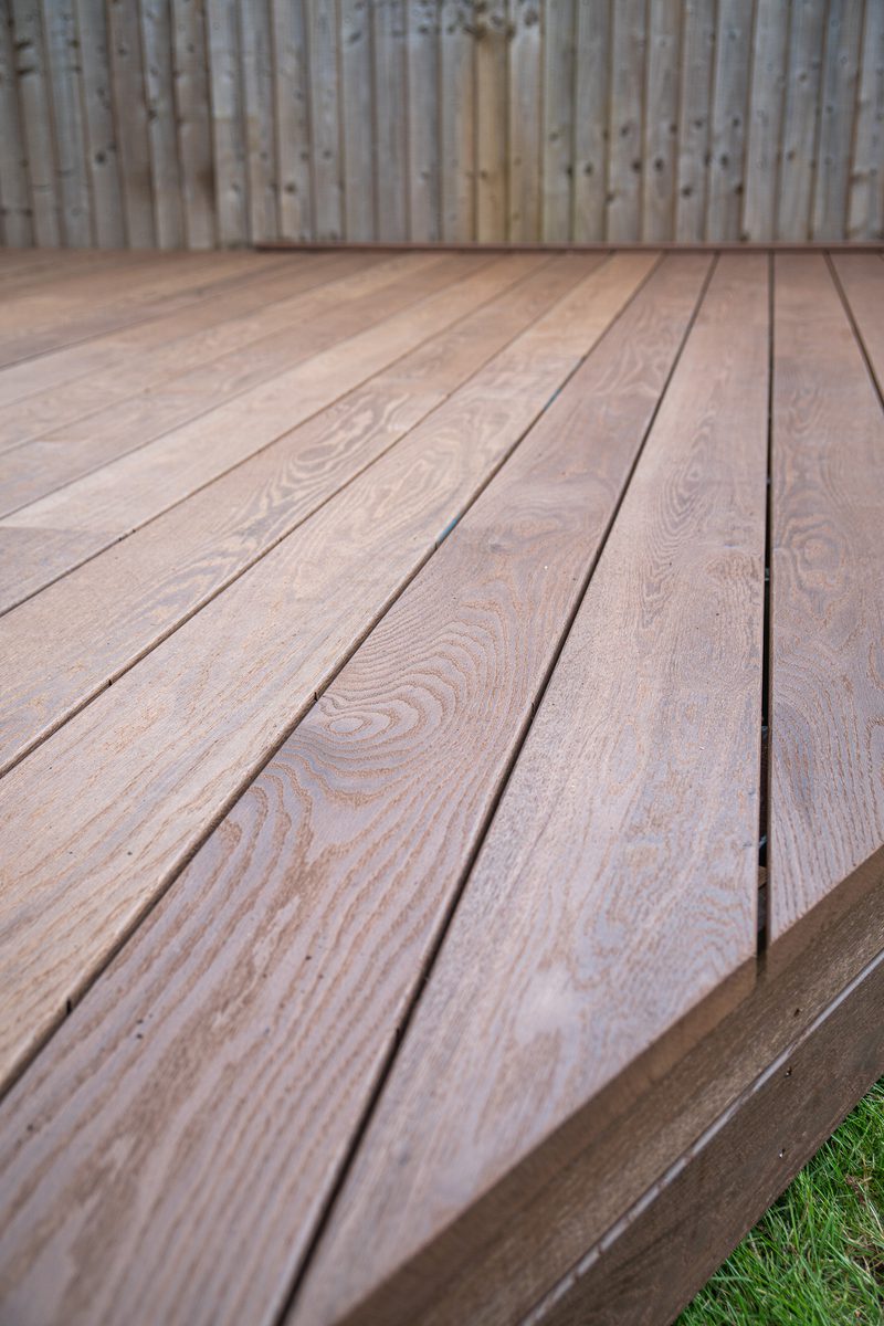 Close-up of a wooden deck with brown planks showing grain, running toward a wooden fence; a strip of green grass appears at the edge.
