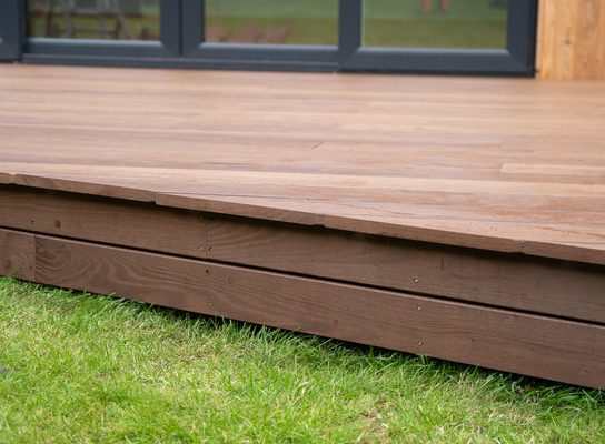Brown wooden deck with visible planks and a raised edge, over a green lawn; dark-framed glass doors and wooden wall visible in the background.