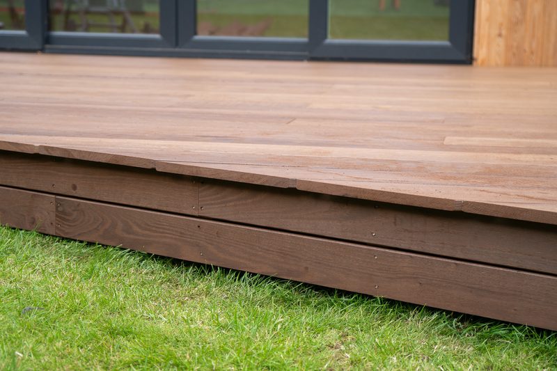 Brown wooden deck with visible planks and a raised edge, over a green lawn; dark-framed glass doors and wooden wall visible in the background.