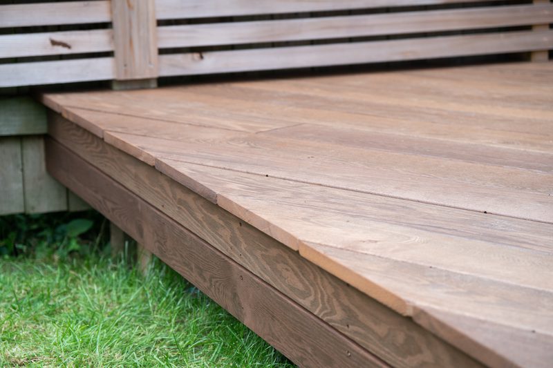 Outdoor wooden deck with wide brown planks and a slatted railing in the background, with green grass beneath.