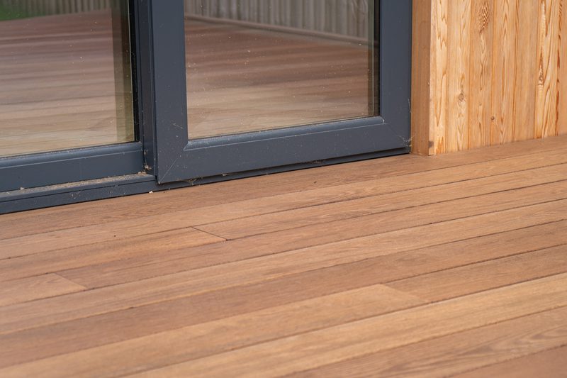 Close-up of a wooden deck with horizontal planks, a dark-framed sliding glass door, and a vertical wooden wall panel on the right.