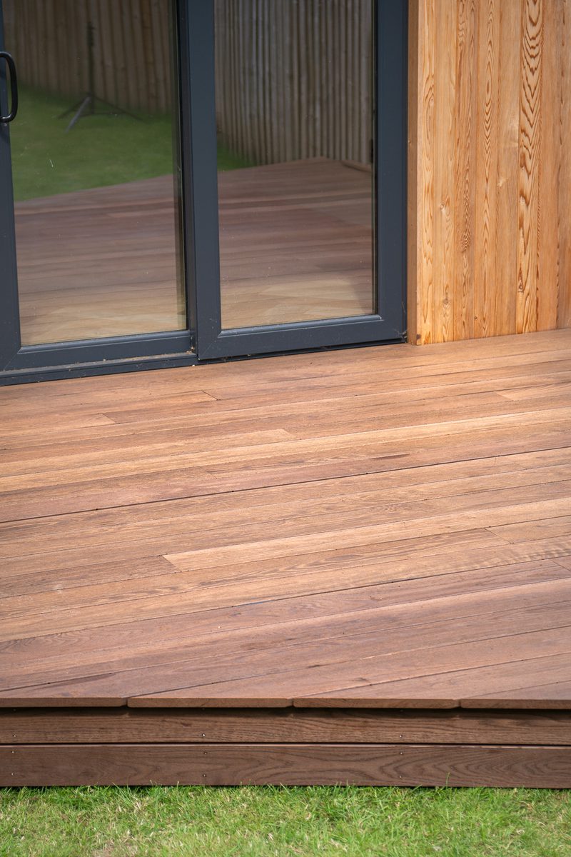 Brown wooden deck with long planks and a glass sliding door with a dark frame, next to a wood-paneled wall; green grass visible at the bottom.