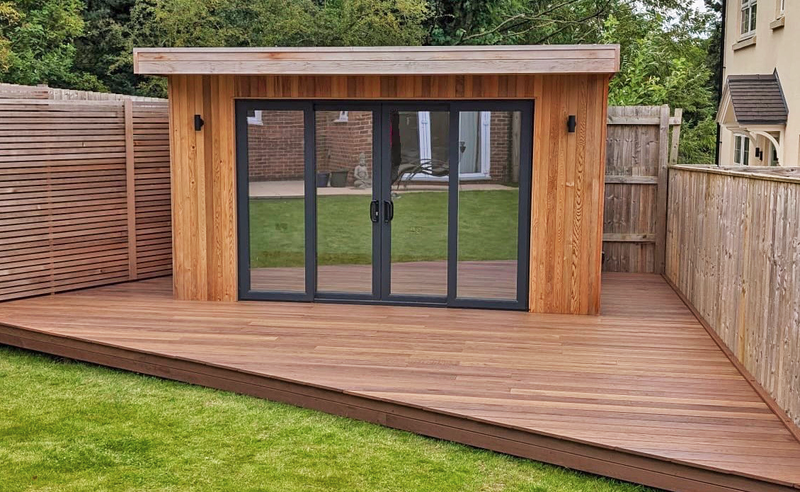 Wooden garden shed with large glass double doors and black frames, built on a wide wooden deck, fenced on both sides with a green lawn behind.