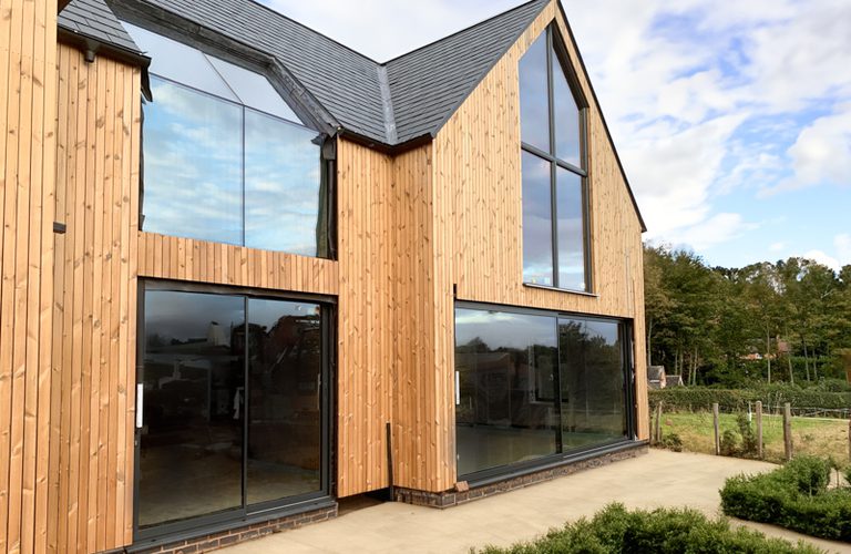 Modern wooden house with tall glass windows and sliding doors, vertical timber cladding and a sloped roof, beside a paved patio and green hedges.