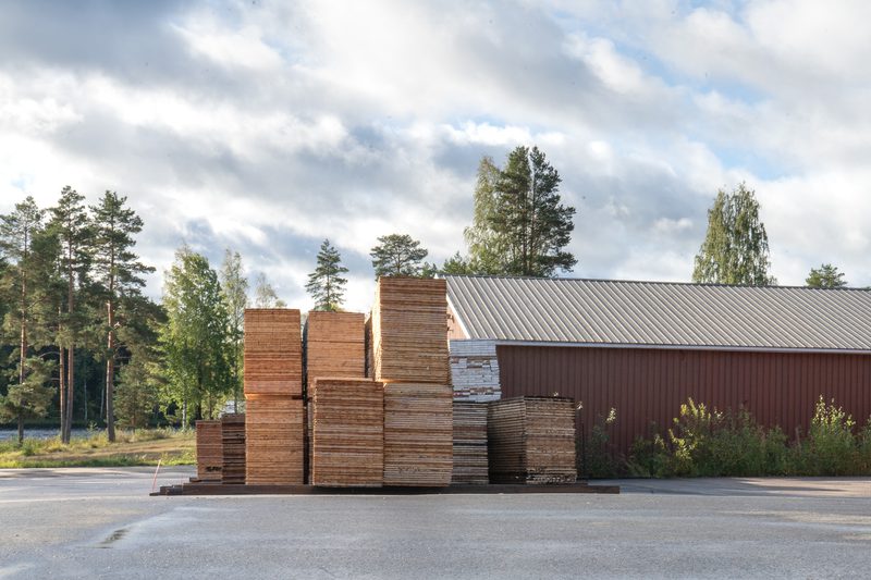 Stacks of lumber planks outdoors beside a red metal-roofed warehouse, with trees and a cloudy sky in the background.