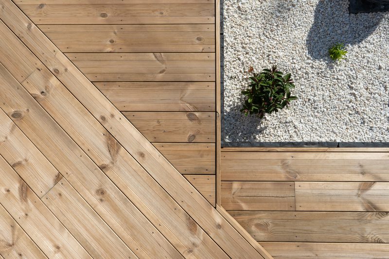 A wooden deck with diagonally laid planks intersects a white gravel bed with a small green shrub; a shadow lies on the gravel near the edge.
