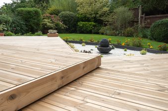 Wooden deck with a stepped platform in a garden, featuring a stone ball fountain on white gravel, orange flowers, and green shrubs.