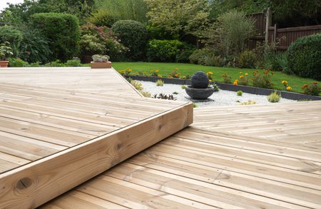 Wooden deck with a stepped platform in a garden, featuring a stone ball fountain on white gravel, orange flowers, and green shrubs.