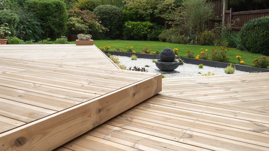 Wooden deck with a stepped platform in a garden, featuring a stone ball fountain on white gravel, orange flowers, and green shrubs.