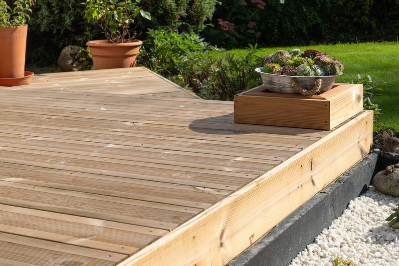 Wooden deck with a metal bowl of succulents on a raised wooden pedestal, flanked by terracotta pots, a green lawn and white gravel.