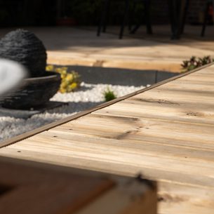 Outdoor wooden deck with diagonal planks, a black stone fountain sphere on white pebbles, blurred foreground pot, and a seating area in background.