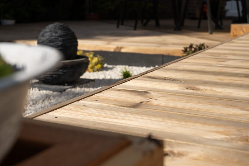 Outdoor wooden deck with diagonal planks, a black stone fountain sphere on white pebbles, blurred foreground pot, and a seating area in background.
