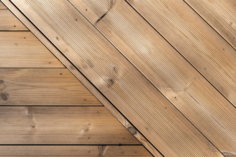 Close-up of light brown wooden deck planks with visible grain, knots, and screws, arranged diagonally across the surface.
