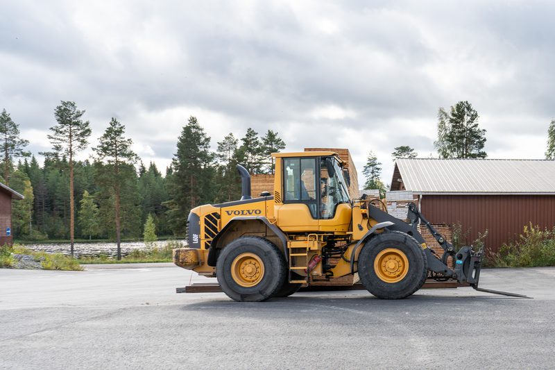 Yellow Volvo wheel loader with pallet forks attached, parked on a paved lot beside wooden buildings, with a forest and cloudy sky in the background.