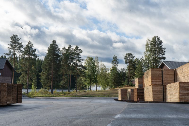 Stacks of wooden planks on pallets at an outdoor lumber yard, with a forest and lake in the background under cloudy skies.