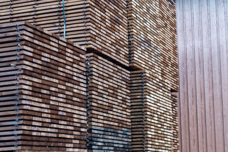 Stacks of brown wooden planks bound in bundles with metal bands; a blue strap hangs among them beside a vertical wooden wall.