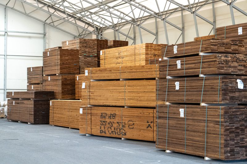 Warehouse interior with tall stacks of brown wooden planks on pallets, strapped and labeled under a metal-framed roof.