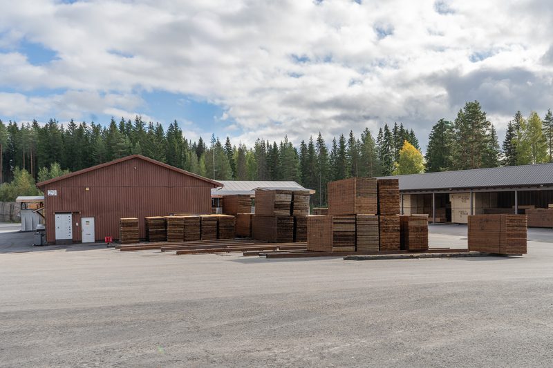 Outdoor lumber yard with stacked wooden boards and pallets, brown sheds, a paved lot, and a forest of evergreens under a cloudy sky.