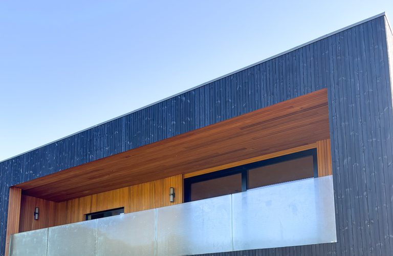 Modern building with dark vertical siding, an open wooden balcony alcove with orange trim and a frosted glass railing under a blue sky.