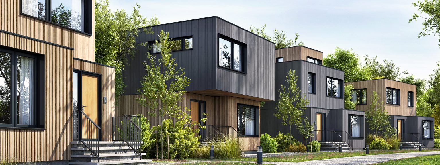 Row of modern two-story houses with wood and dark gray cladding, large windows, stairs to front doors, and lush landscaping along a gravel path.