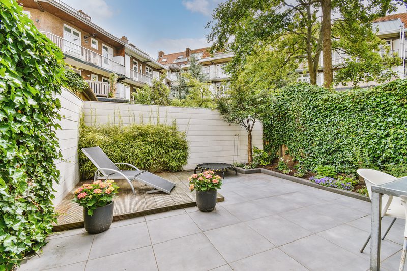 Urban courtyard patio with gray tiles, a sun lounger on a wooden deck, potted flowers, ivy-covered walls, a tree, and surrounding apartment buildings.