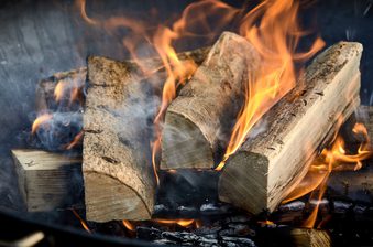 Split wooden logs burning in a fireplace, bright orange flames curling around the wood with wisps of smoke and glowing embers below.