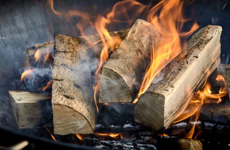 Split wooden logs burning in a fireplace, bright orange flames curling around the wood with wisps of smoke and glowing embers below.