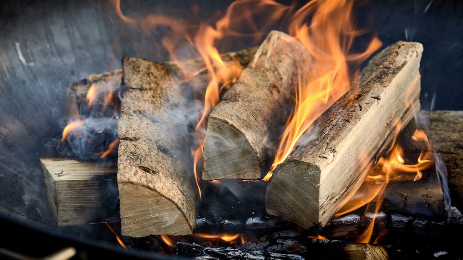 Split wooden logs burning in a fireplace, bright orange flames curling around the wood with wisps of smoke and glowing embers below.