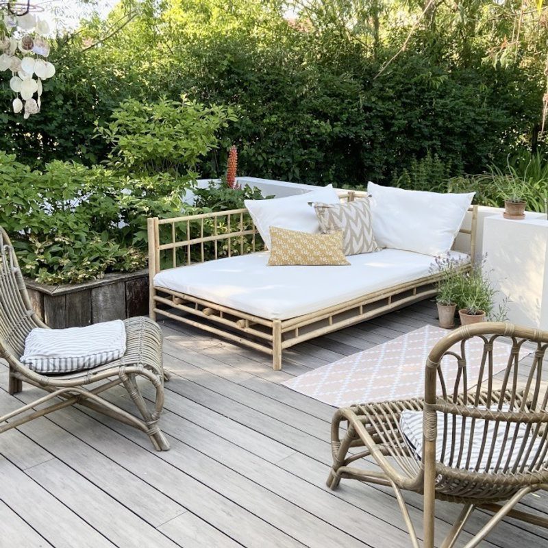 Outdoor wooden deck with a bamboo-framed daybed, two wicker chairs, white cushions, patterned pillows, and potted plants along a green hedge.