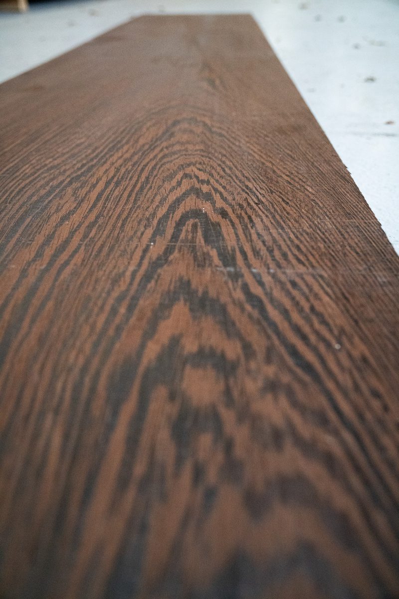 Close-up of a long dark brown wooden plank with prominent grain patterns, extending away toward a light background.