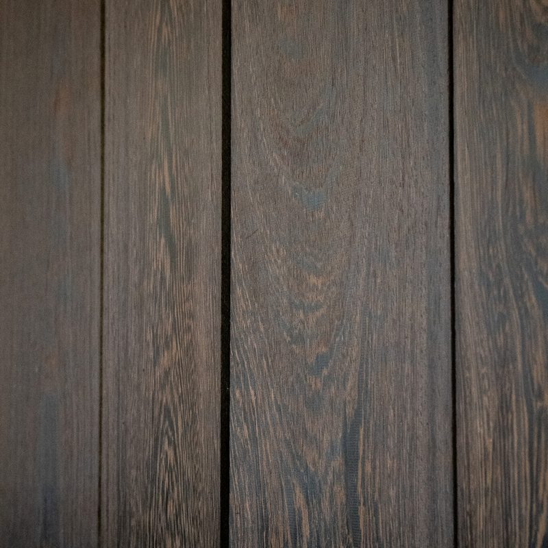 Close-up of vertical dark brown wooden planks with prominent grain and narrow gaps; a lighter blurred wooden edge appears in the foreground.
