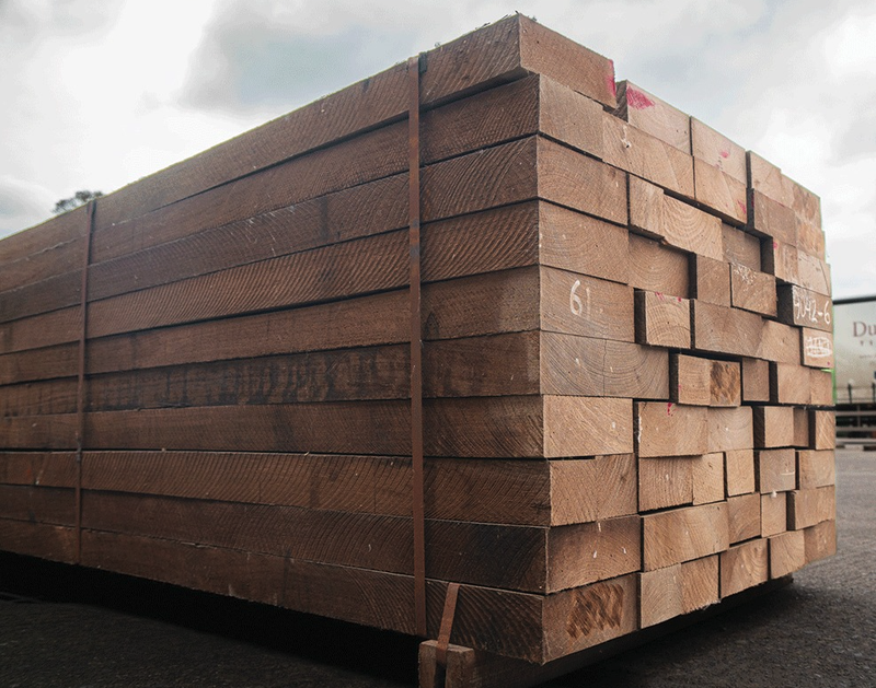Large pallet stacked with brown wooden planks bound by straps, outdoors on asphalt under a cloudy sky.