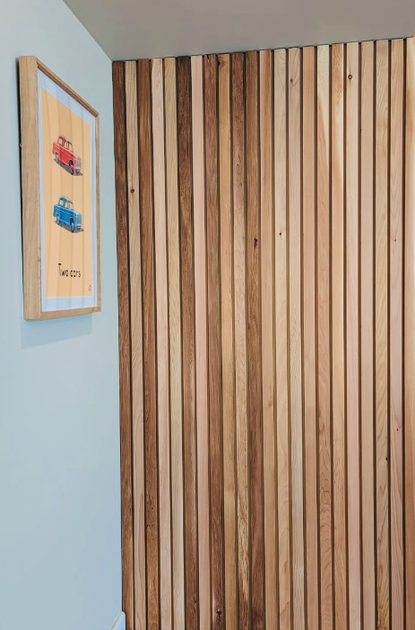 Corner of a room with a light blue wall, a framed poster of two cars, and a vertical wooden slat wall; wooden floor and a ceiling spotlight.