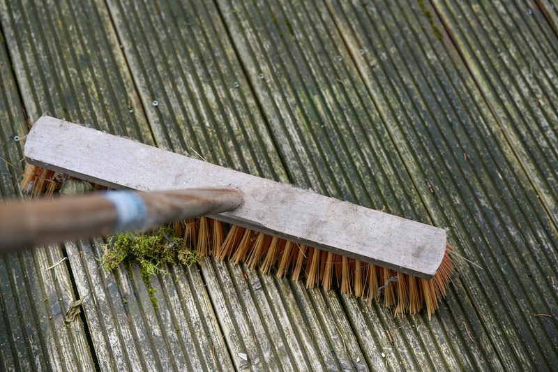A broom with a long wooden handle and brown bristles rests on a weathered wooden deck with moss and dirt between the boards.